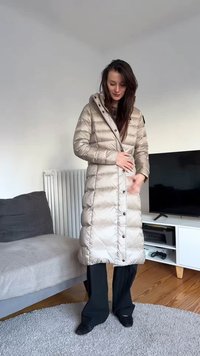 Beige dunjakke med lodret quiltet mønster, knapper foran og hætte. Båret over sorte bukser med vide ben, der har en blank, glat tekstur.