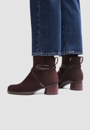 Mørkebrune ankelstøvler med lynlås i siden, dekorativt bånd og spænde, båret med blå cropped jeans mod en ensfarvet lys baggrund.