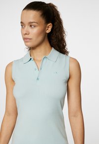 En ärmlös ljusblå polo-tröja i mjukt stickat material, med krage, tre knappar och sidodetaljer i mesh-textur.