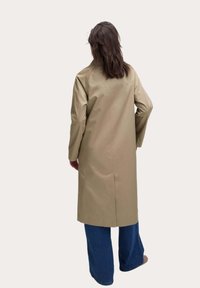 Beige waterdichte trenchcoat met lange mouwen en een rechte snit, gedragen over wijde denim broeken. De achterkant heeft een middennaad.