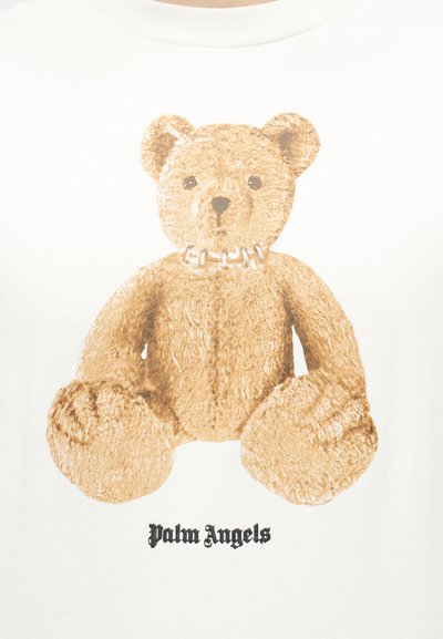 Ours en peluche marron portant un collier chaîne argenté, centré sur un fond blanc avec le texte "Palm Angels" en dessous.