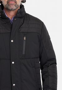 Chaqueta acolchada negra con cuello alto, paneles cosidos horizontalmente, bolsillo en el pecho con cremallera y cierre a presión; el tejido parece suave y mate.