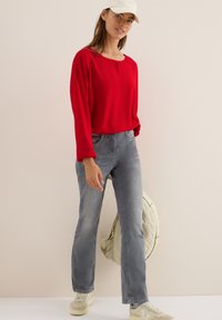 Röd långärmad blus med rund halsringning, i kombination med ljusgrå jeans. Modellen bär en vit keps och krämfärgade sneakers.
