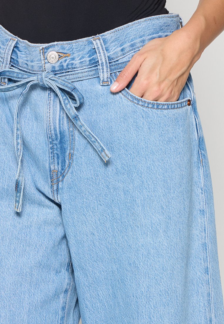 Jean en denim bleu clair avec taille haute, ceinture à nouer et poches avant. Présente une texture délavée et des détails de couture contrastants.