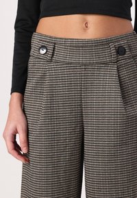 Bruine en zwarte houndstooth-patroonbroek met twee voordoekjes, twee zwarte knopen en een trouw aansluitende pasvorm bij de taille.