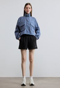 Κοντό μπλε μπουφάν με δύο μπροστινές τσέπες, ελαστικά μανίκια, φορεμένο πάνω από μαύρη βερμούδα και λευκές κάλτσες, συνδυασμένο με ανοιχτόχρωμα αθλητικά παπούτσια.