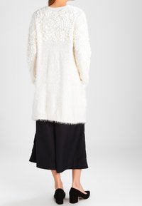 Lange witte fluffy cardigan met textuur van bloemaccenten op de achterkant, gecombineerd met zwarte culottes en gesloten zwarte hakken.