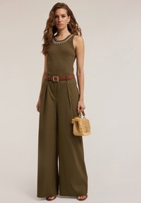Olijfgroene mouwloze top met decoratieve kralen bij de halslijn, gecombineerd met hooggelegde, wijde broek en een geweven handtas.