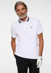 Biała koszulka polo z czarno-białym paskowanym kołnierzykiem oraz małym czarnym logo na lewej piersi. Widoczne czarne spodnie i biała rękawica golfowa.