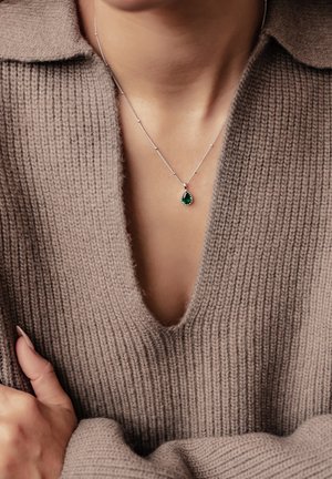 Silver necklace with a pear-shaped green pendant, worn over a soft taupe knit sweater featuring a wide V-neck.