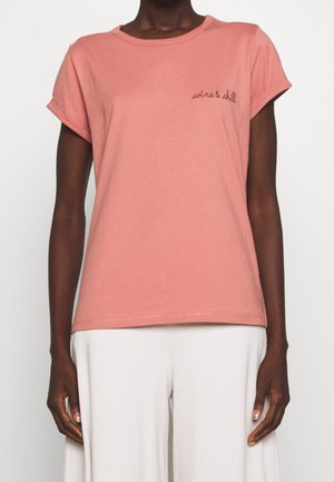 Personne portant une chemise rose délavée à manches courtes avec l'inscription « wine & chill » sur la poitrine, associée à un pantalon blanc ample.