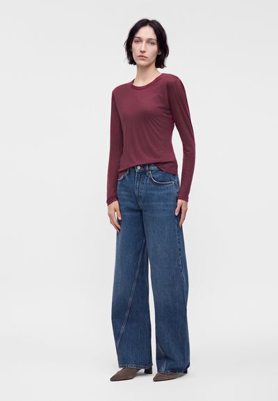 Femme aux cheveux courts et foncés portant un haut bordeaux ajusté à manches longues, un jean bleu à jambes larges et des chaussures marron pointues, debout en pose neutre.