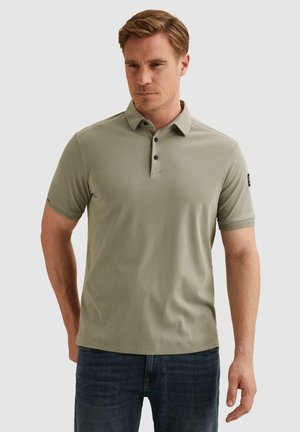 Homme portant un polo vert clair à manches courtes et un jean bleu foncé, debout devant un fond clair uni.