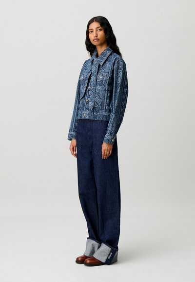 Donna che indossa una giacca di jeans blu a fantasia, jeans a gamba larga blu scuro con grandi risvolti e scarpe marroni, in piedi su uno sfondo neutro.