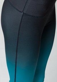 Legginsy w gradientowym wzorze przechodzącym od ciemnoszarego u góry do szmaragdowego u dołu, wykonane z gładkiego, elastycznego materiału z płaskimi szwami.