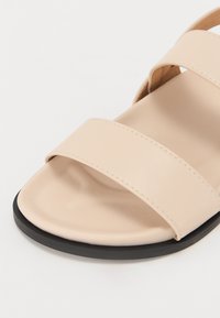 Sandale en synthétique beige avec deux larges sangles, un dessus lisse et une semelle en caoutchouc noir avec un design de bout arrondi.