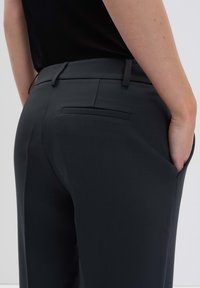 Pantalons gris foncé avec une texture lisse, dotés d'une poche arrière et de passants de ceinture, présentés de côté avec une main dans la poche.
