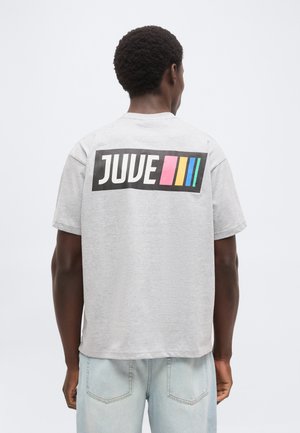 Homme aux cheveux courts et bouclés porte un t-shirt gris clair avec "JUVE" et des bandes colorées en rose, jaune et bleu sur le haut du dos, jean clair.
