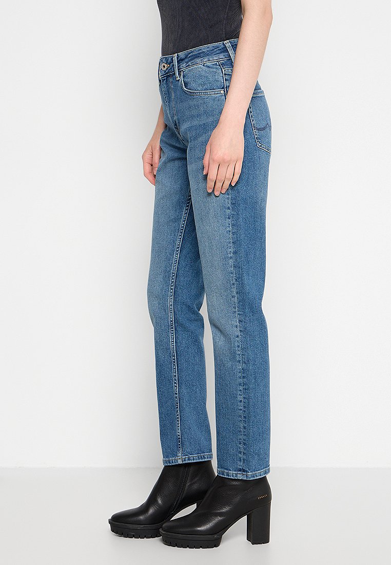 Frau trägt hoch taillierte, gerade geschnittene blaue Jeans mit schwarzen, schulterfreien Ankle Boots vor weißem Hintergrund.