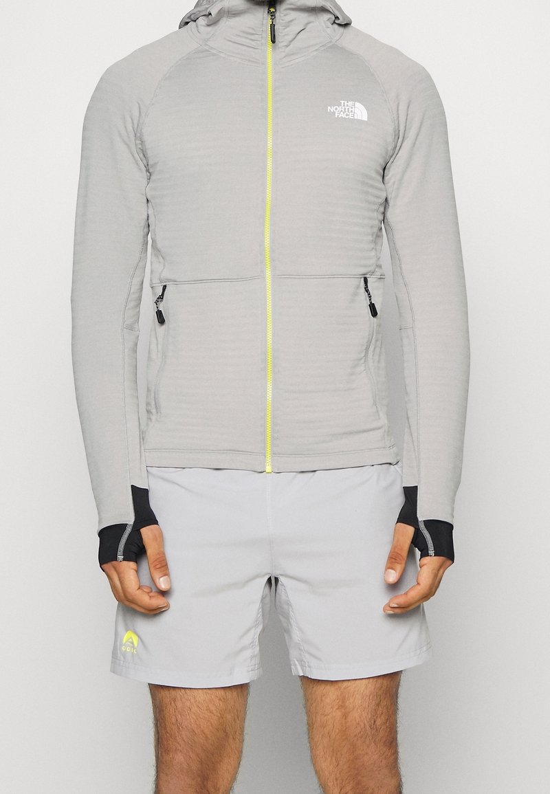 Homme portant une veste à capuche gris clair avec fermeture éclair et trous pour les pouces et un short de sport gris clair avec un logo sur la jambe gauche, debout devant un fond blanc.