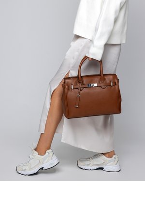 Person wearing white sneakers and a light skirt with a side slit, carrying a large brown leather handbag by its handles.