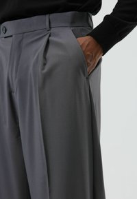 Pantalon gris plissé avec une texture lisse, une taille ajustée et des poches latérales. Le tissu semble léger et épuré.