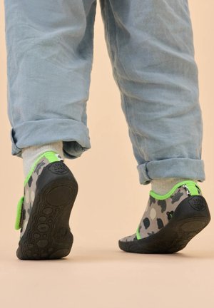 Enfant portant un pantalon gris et des chaussures à motif avec une bordure vert vif, marchant loin sur une surface beige.