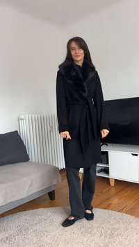 Casaco longo preto com gola de pele sintética, detalhe de cintura ajustada, usado sobre calças escuras, combinado com sapatos planos pretos, num ambiente luminoso.