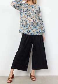 Blusa fluida e stampata in beige con design floreale blu e nero, abbinata a pantaloni neri a gamba larga plissettati e sandali con tacco a blocco marroni.
