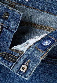 Nærbilde av blå denimjeans med åpnet glidelås som viser glidelås, knapp, justerbar elastisk midje og hvit indre klesetikett.