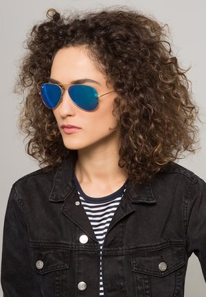 Occhiali da sole aviator con lenti blu specchiate, sottili montature in metallo dorato, indossati con una giacca in denim nera e una camicia a righe.