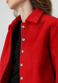 Rode wollen jas met een kraag, voorzien van zes ronde knopen aan de voorkant en een textuurrijke afwerking. Draagt over een zwart shirt.