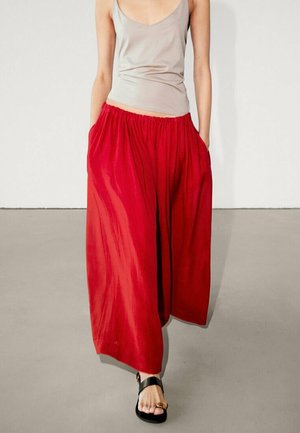 Femme portant un pantalon rouge ample à jambes larges, un haut beige clair sans manches et des sandales noires à bout ouvert, marchant sur un sol gris devant un mur blanc.