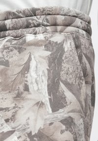 Kamuhelyes melegítőnadrág, texturált anyaggal, szürke és földszínek keverékével, levél- és fakéreg mintázattal. Rugalmas derekú.