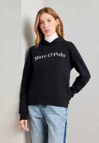 Schwarzer Kapuzenpullover mit einem Frontlogo, das "Marc O'Polo" liest. Mit einer Kordelzugkapuze und gerippten Bündchen; getragen über hellblauen Jeans.