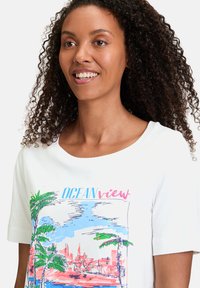 Hvid bomulds T-shirt med korte ærmer, prydet med et farverigt trykt design af palmetræer og en havudsigt. Rund halsudskæring, afslappet pasform.