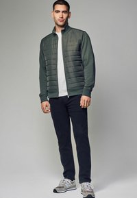 Grüne Zip-Jacke mit gestepptem Vorderteil, graues T-Shirt, dunkle Hose und graue Sneakers. Hat gerippte Bündchen und Saum.