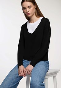 Maglione nero lavorato a maglia con scollatura a V, indossato sopra una camicia bianca. Il modello è seduto su uno sgabello bianco, abbinato a jeans in denim azzurro chiaro.