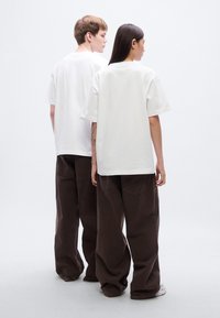 Δύο άτομα φορούν υπερμεγέθη λευκά μπλουζάκια και χαλαρά καφέ παντελόνια, με απλό σχέδιο και χαλαρή γραμμή, εμφανιζόμενα από πίσω.