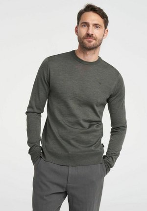 Dunkelgrüner Langarmpullover aus einem weichen Material, mit einem Rundhalsausschnitt und dezentem Logodetail. Kombiniert mit grauen Hosen.
