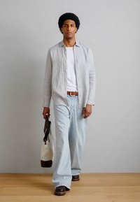 Άνδρας που περπατάει μέσα σε χώρο φορώντας ανοιχτό γκρι πουκάμισο πάνω από λευκό μπλουζάκι, ανοιχτό μπλε φαρδιά τζιν παντελόνια, καφέ παπούτσια, κρατώντας τσάντα ώμου μπεζ και καφέ.