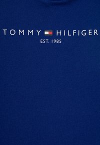 Modrá bavlněná tričko s bílým textem, který říká "TOMMY HILFIGER EST. 1985", s malým barevným logem mezi slovy. Hladká textura.