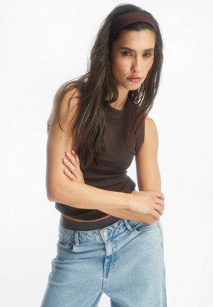 Młoda kobieta o długich ciemnych włosach, ubrana w ciemny bezrękawnik, niebieskie dżinsy i pasującą opaskę na głowę, skrzyżowanymi ramionami i neutralnym wyrazem twarzy.