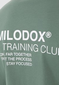 Grøn bomulds T-shirt med fed hvid tekst der står "MILODOX® TRAINING CLUB" og inspirerende sætninger. Blød, glat tekstur.