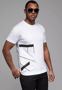 Hvid bomulds t-shirt med et sort abstrakt design på forsiden. Bæres med sorte bukser og solbriller. Strukturert stof, rund hals.