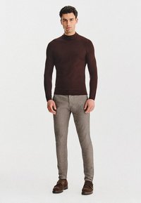 Brązowy sweter z długim rękawem w połączeniu z jasnoszarymi, dopasowanymi spodniami i brązowym skórzanym obuwiem. Ubranie ma gładką teksturę i dopasowany krój.