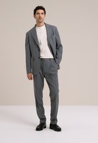 Grauer Nadelstreifenanzug mit strukturiertem Blazer und gerade geschnittenen Hosen, kombiniert mit einem weißen T-Shirt. Schwarze Schuhe runden den Look ab.