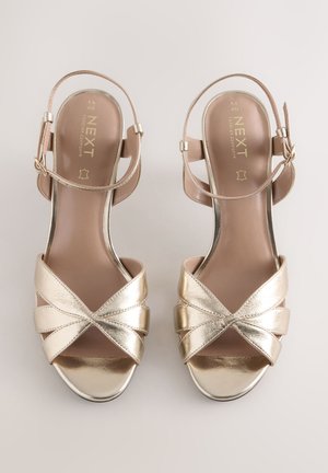 Sandales à talons métalliques dorées à deux lanières avec une conception à bande croisée, une bride de cheville réglable et une semelle intérieure lisse beige clair.