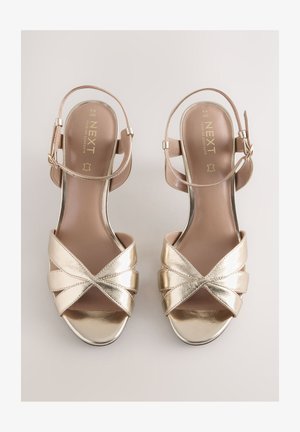 Kultaiset metallinhohtoiset korkosandaalit, joissa on kaksi hihnaa ja ristiin aseteltu nauha, säädettävä nilkkahihna sekä sileä, vaaleanbeige sisäpohja.