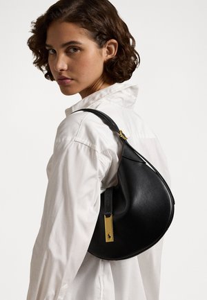 Bolso de hombro de cuero negro con forma curva, que presenta un acento de hardware dorado y una textura suave. Llevado sobre una camisa blanca.